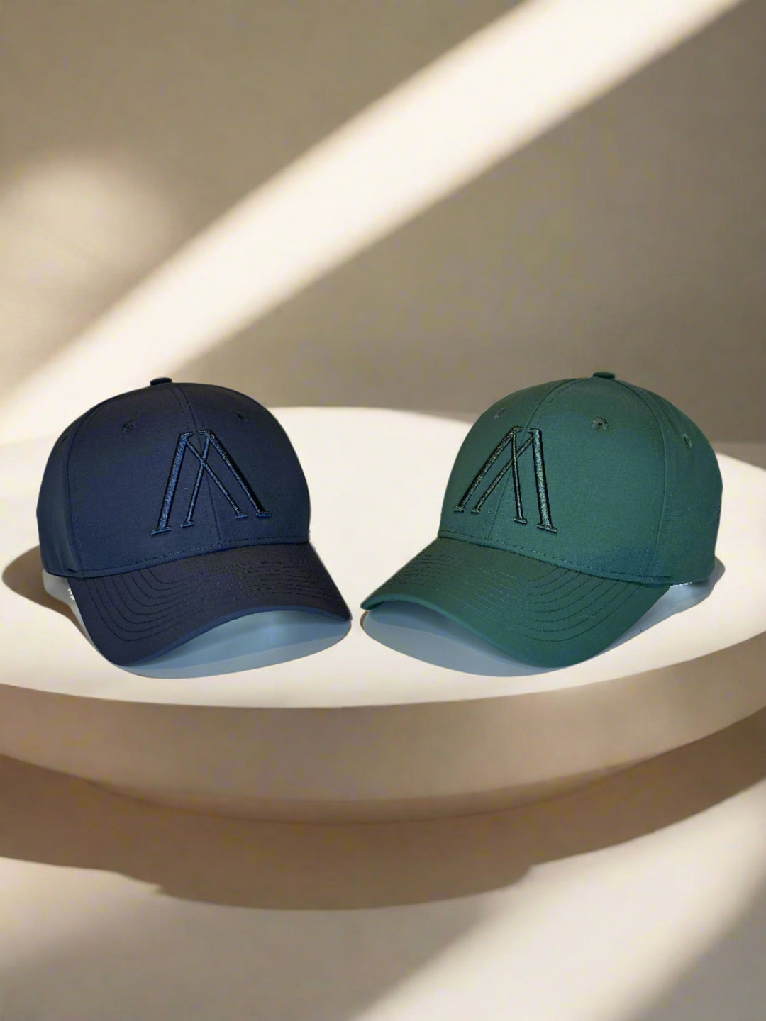 Zwei Baseballkappen auf Podest: dunkelblau mit A-Logo, grün daneben