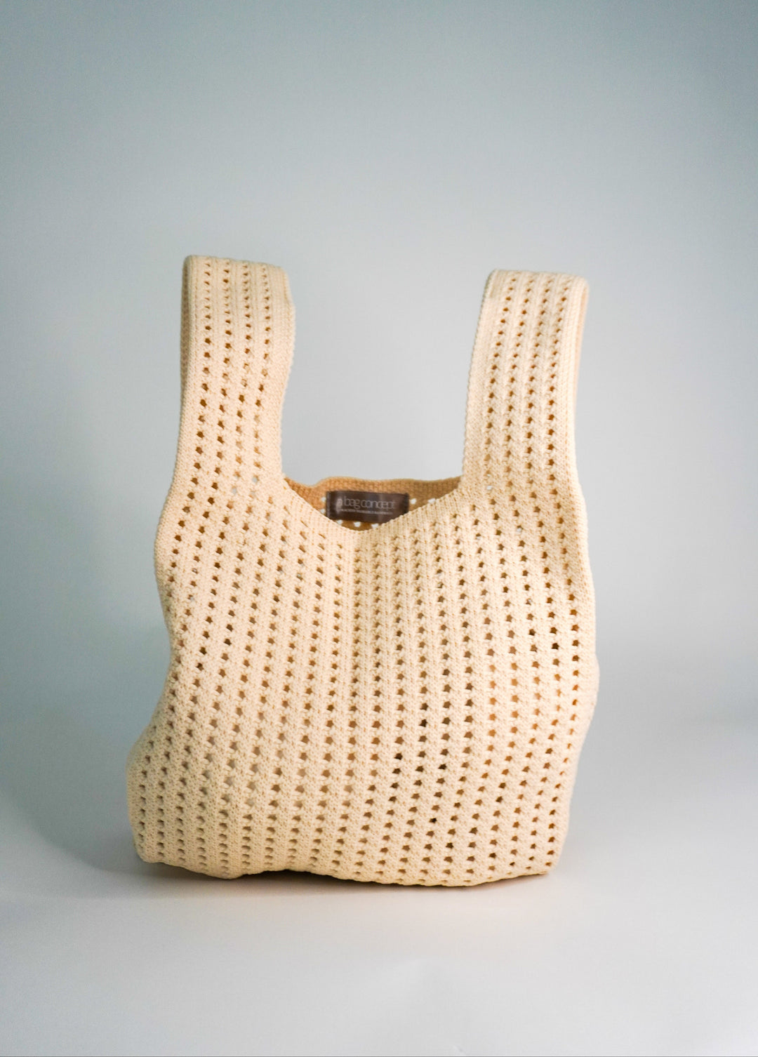 Oat crochet drawstring handbag, front view with gathered neck opening and small inner tag.