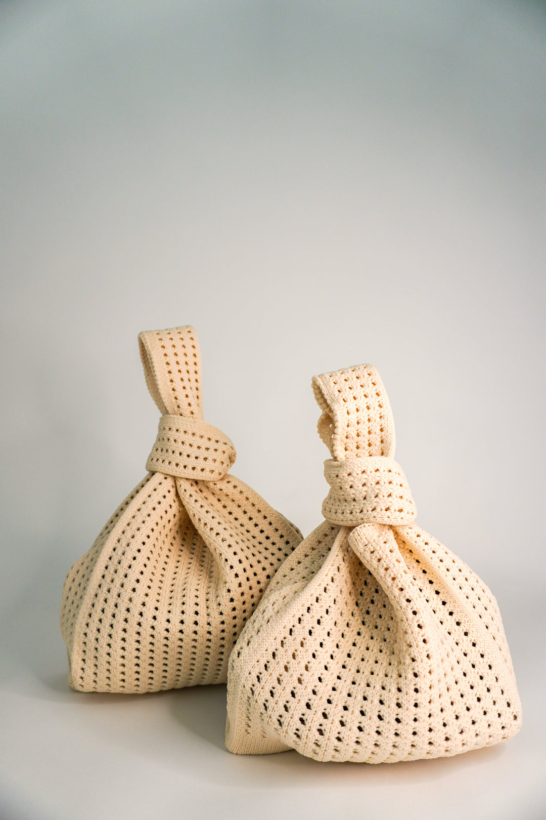 Pair of oat crochet drawstring handbags with knotted top ties and openwork lattice texture.
