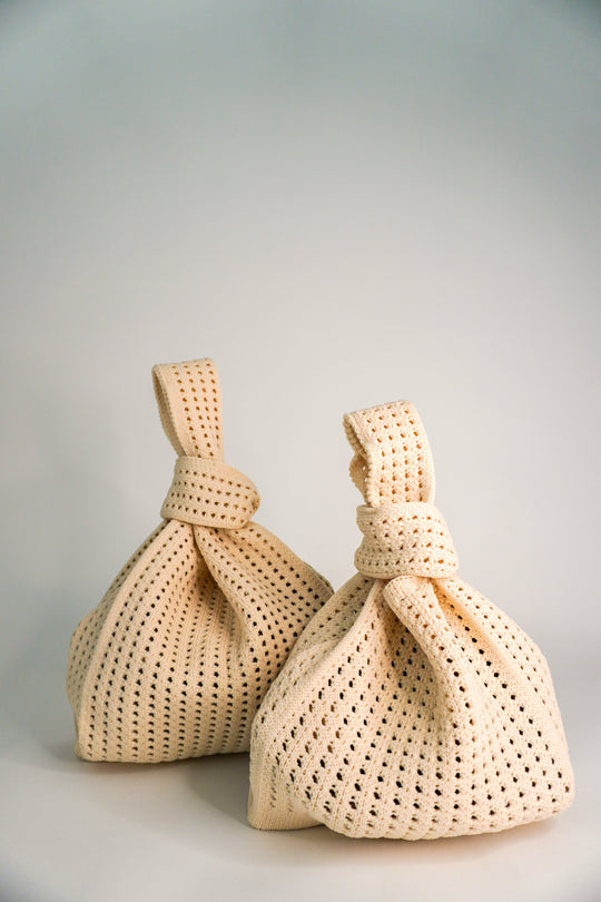 Pair of oat crochet drawstring handbags with knotted top ties and openwork lattice texture.