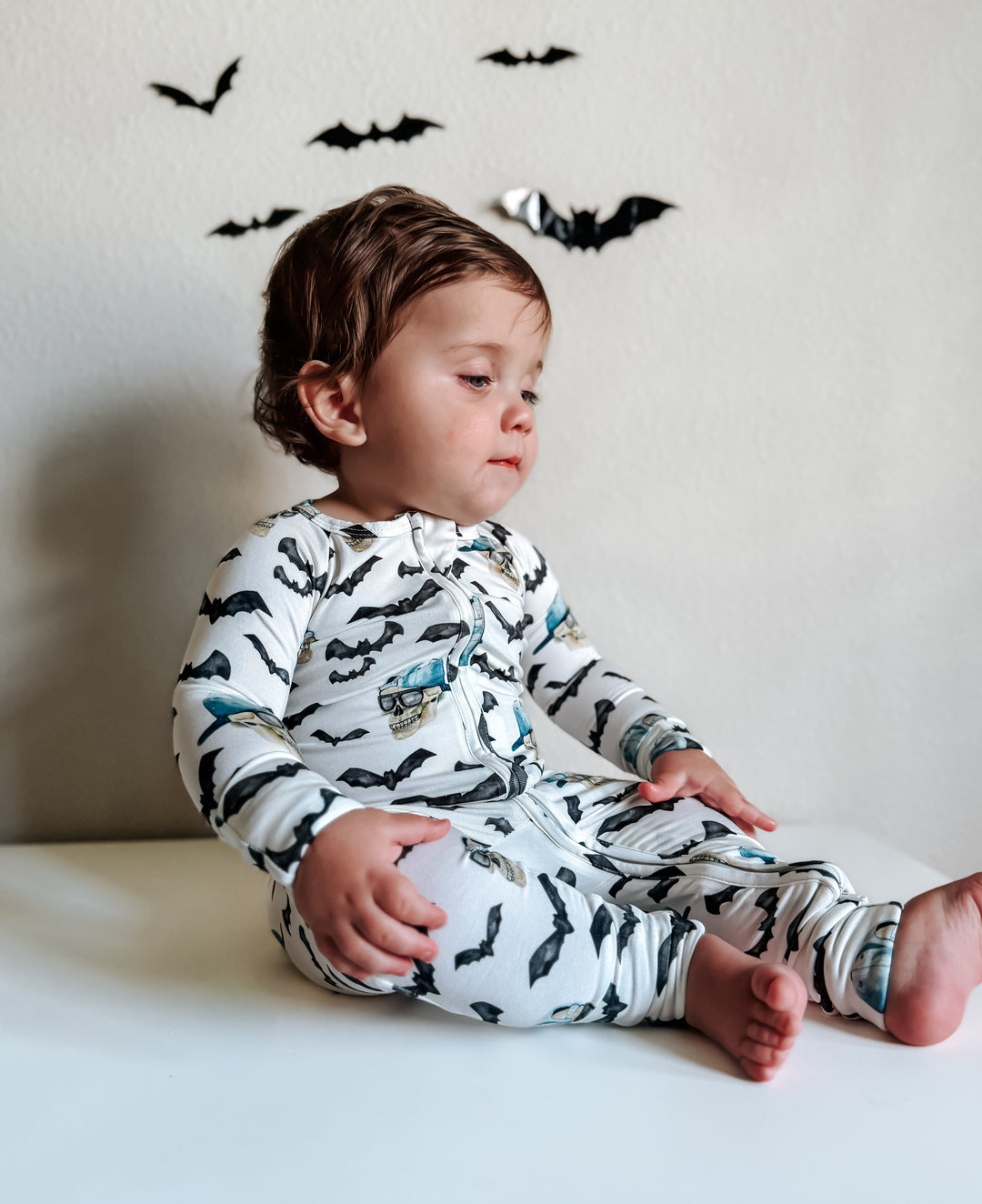 Child seated wearing zippy pajamas; white fabric with black bats and blue skulls, zip closure