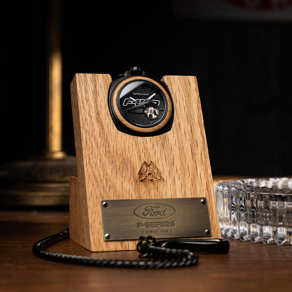 Reloj de bolsillo vintage sobre base de madera con placa metálica y cadena negra.