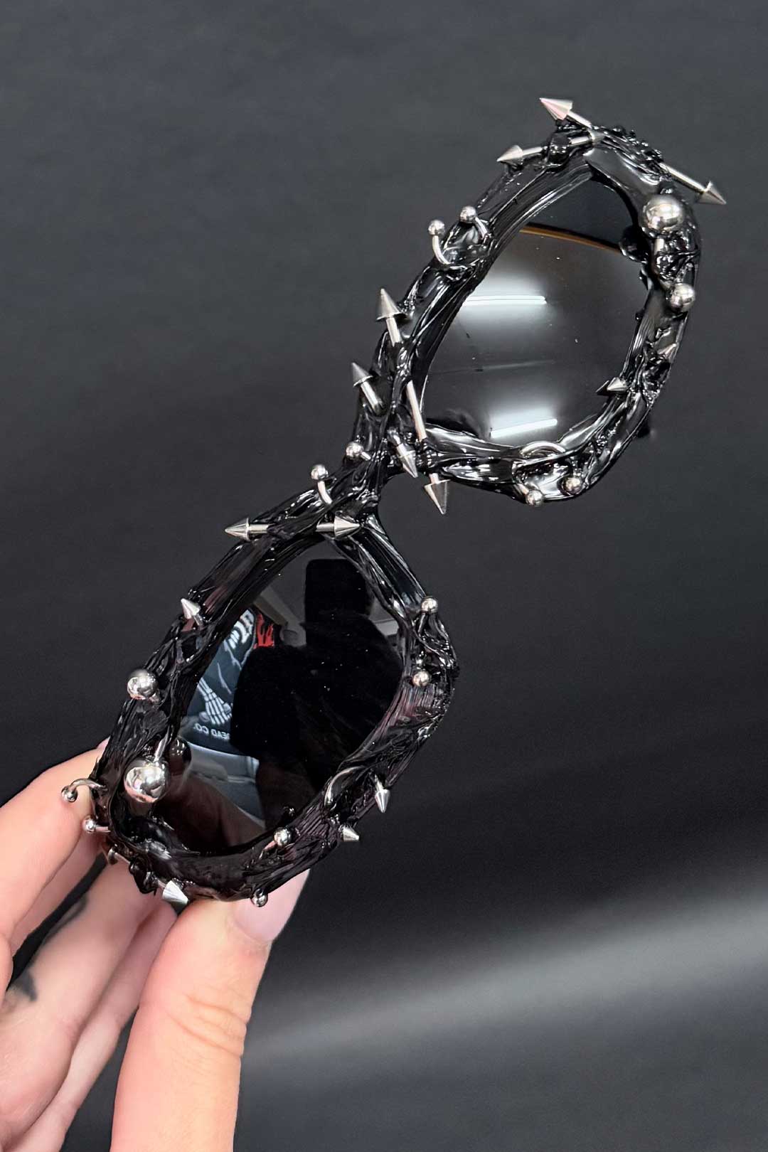 Holy Sunglasses with black lenses and chrome spiked chain frame, held in hand