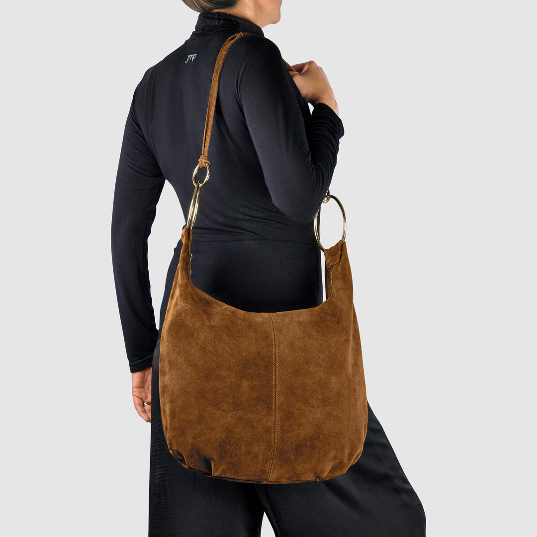 Brown suede hobo bag worn on shoulder with gold ring handles and rounded open top