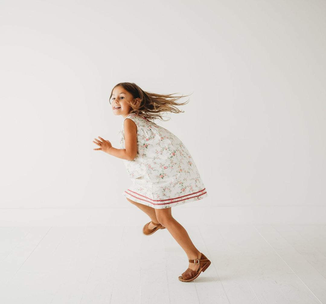 Girl wearing white rosebud cotton tea dress with burgundy hem trim and sleeveless cut